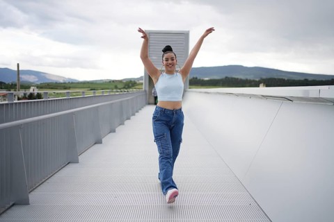 A woman in blue jeans and a blue top is dancing on a bridge. The bridge is made of metal and is