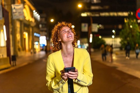Happy woman looking up while strolling along the city streets using mobile phone at night
