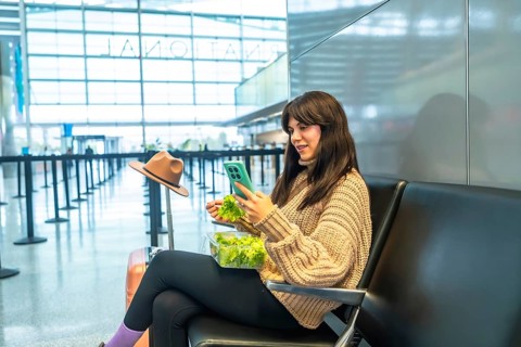Young woman sitting in a modern airport terminal, eating a fresh green salad from a plastic