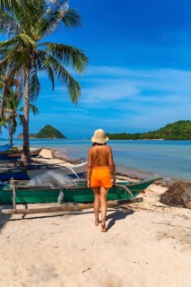 Young woman walking on a white sand beach next to traditional fishing boats, enjoying a sunny day