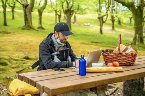 A young Caucasian father teleworking sitting at a picnic table with computer in the woods. Summer