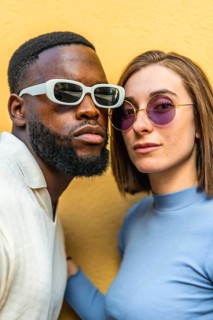 Vertical close-up photo of a stylish multiracial couple wearing sunglasses looking at camera