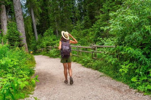 Female tourist with straw hat and backpack walking on gravel path in forest near lake carezza,