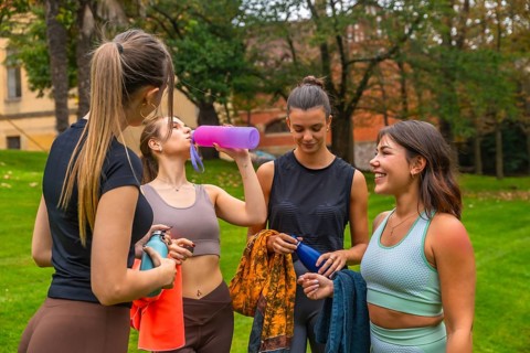 Four young women taking a break and hydrating with reusable water bottles, conversing and smiling