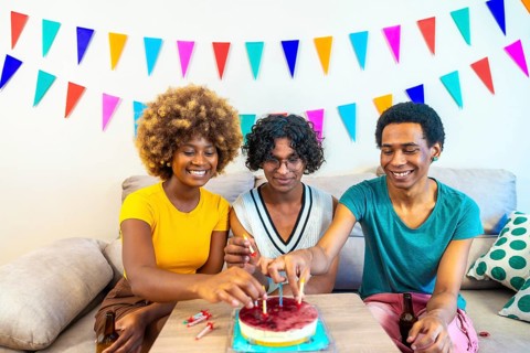 Frontal photo of three multi-ethnic friends placing candles on birthday cake sitting on the sofa