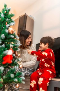 Mother and son in pajamas laughing as they decorate a small christmas tree at home, sharing cozy,