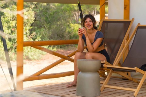 Young woman relaxing on a wooden patio of a glamping tent, savoring a refreshing drink while