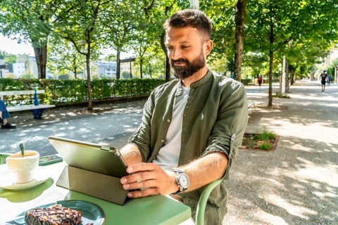 Bearded man at an outdoor cafe working on a tablet, enjoying coffee and cake in a sunny parklike