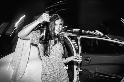 Happy young woman exiting a luxury limousine car, holding a champagne bottle and glass, celebrating