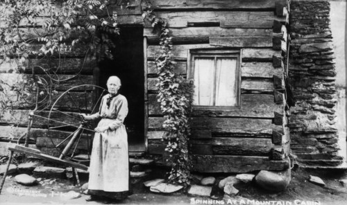 Woman with Spinning Wheel Beside Log Cabin, circa 1910