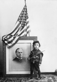 Boy in Sailor Uniform holding Sword in front of flag-draped, head-and-shoulders portrait of William McKinley, 1896