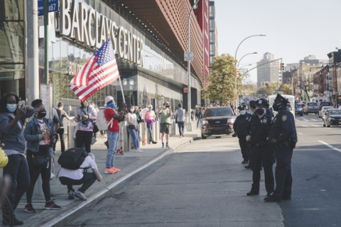 Man holding up American Flag at Barclays Center, opposite a Line of Police during Celebration of President-Elect Joe Biden