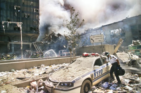 Rescue worker reaching into a New York Police car covered with debris with smoldering ruins in background