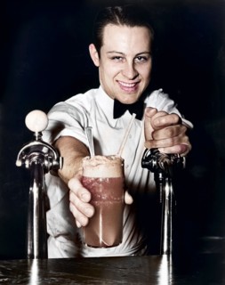 Soda jerk passing ice cream soda between two soda fountains