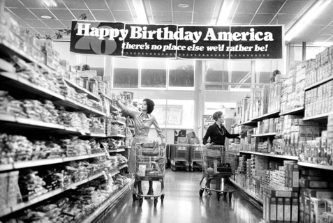 Two women shopping in grocery store under banner reading: "Happy birthday America, there's no place