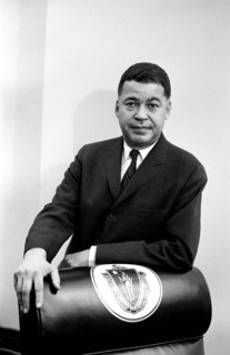 Senator Edward Brooke standing behind chair decorated with Great Seal of the Commonwealth of