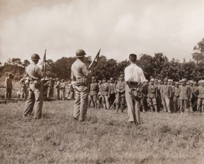 Japanese soldiers line up for formal surrender ceremonies as U.S. Marines with tommy guns and fixed