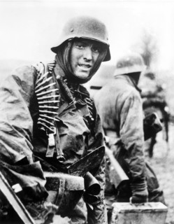 Nazi soldier, heavily armed, carrying ammunition boxes forward with companion in territory taken by