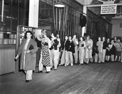 Women war workers wearing slacks punching time cards at end of shift, De Soto bomber plant,