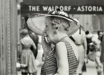 Half-length portrait of elderly woman in wide-brimmed hat standing outside Waldorf-Astoria Hotel,