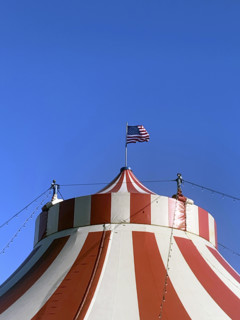 Top of red and white circus tent with American flag against blue sky