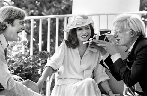 American artist Andy Warhol taking Polaroid picture while sitting with Jack Ford and Bianca Jagger