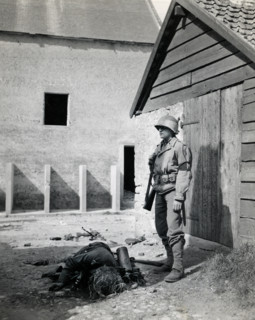 U.S. military police standing guard in barnyard near body of dead German sniper, Normandy, France,