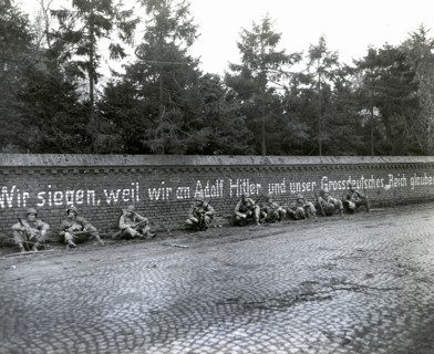 Members of 16th regiment, 1st Division, U.S. First Army, sitting under German slogan "Wir Siegen,