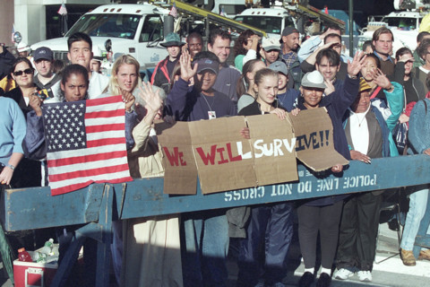 Onlookers waving and holding American flag and sign reading "We Will Survive" during U.S. President