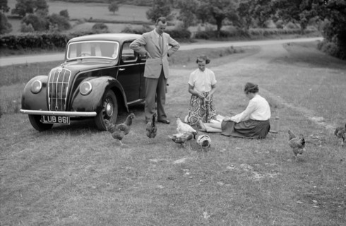A family group feed chickens beside their car, c1945-c1965