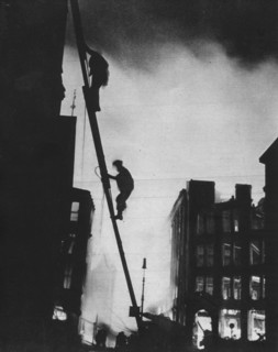'Out in the Blitz, silhouetted aganinst the light of fires, rescue men climb into a building in St.  Artist: Unknown.