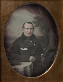 Man Wearing Coat and Gloves, Holding Hat, Seated in Front of Painted Outdoor Backdrop, 1840s.