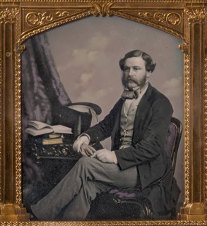 Man Seated in Front of Cloud Backdrop, Books and Top Hat on Table, 1850s.