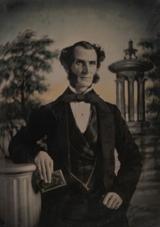Man Holding Book, Standing Before a Painted Scenic Backdrop, 1850s-60s.