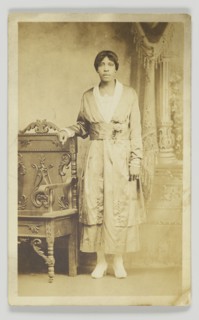 Photographic postcard of a woman standing next to a wooden chair, 1904-1918. Creator: Unknown.