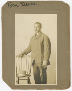 Photograph of Tom Durr standing next to a chair with a hat on it, ca. 1910. Creator: Unknown.