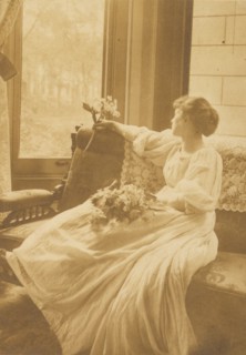 Young woman seated by a window with flowers in her lap, c1900.