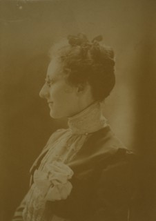 Studio portrait of young woman in white dress posed as if holding flowering tree branch, c1900.