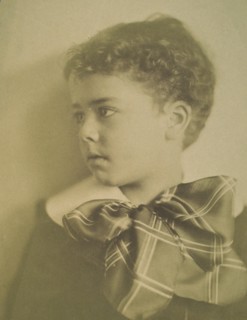 Head-and-shoulders portrait of a young boy with a large plaid bow at the neck of his jacket, facing left, c1900.