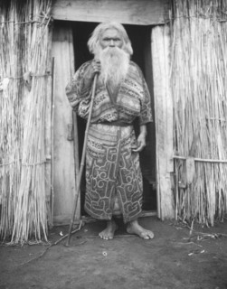 Ainu man holding a staff standing at the doorway of a hut, 1908. Creator: Arnold Genthe.