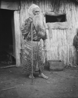 Ainu man holding a staff standing outside a hut, 1908. Creator: Arnold Genthe.