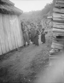 Group of Ainu children standing in a passageway between huts, 1908. Creator: Arnold Genthe.