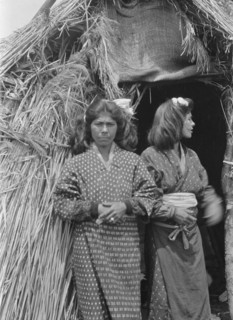 Two Ainu women standing outside the entrance of a hut, 1908. Creator: Arnold Genthe.
