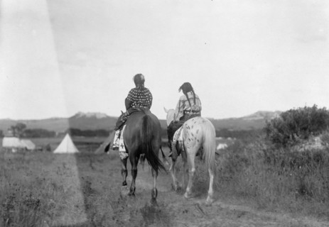 Two daughters of a chief on horseback, riding away from camera toward tents in background, c1907. Creator: Edward Sheriff Curtis.