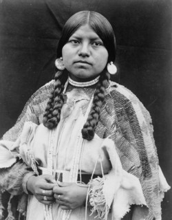 Cayuse woman, half-length portrait, standing, facing front, braids, shell disk earrings..., c1910. Creator: Edward Sheriff Curtis.