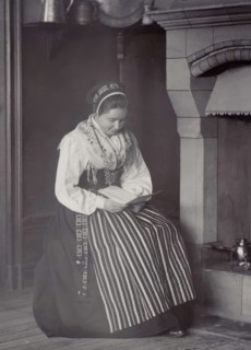 A woman sits in front of a fireplace and reads dressed in a Dalarna costume, 1886-1920. Creator: Helene Edlund.