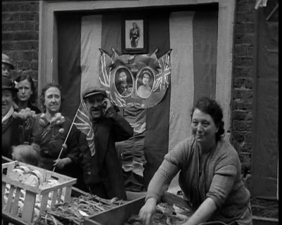 Woman and Others Standing Behind a Street Stall With Wall Behind Decorated With Flags..., 1937. Creator: British Pathe Ltd.