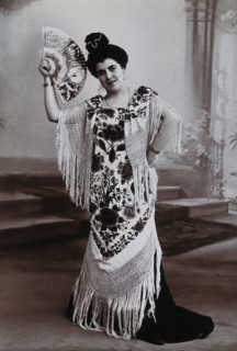 A standing woman, holding aloft a fan, wearing a fringed shawl, in front of a painted..., c1900. Creator: Unknown.