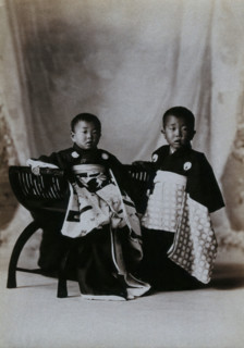 Two boys in a photographic studio, one seated on a chair, in front of a curtained backdrop, c1900. Creator: Unknown.