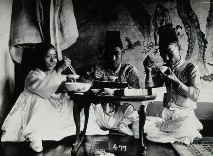 A group of people eating a meal, seated behind a small table, in front of a painted screen, c1900. Creator: Unknown.